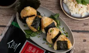 Pięć onigiri z ryżem, udekorowanych listkami nori i szczypiorkiem, obok opakowania nori i miseczki z ryżem.