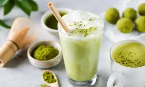 Orzeźwiający iced matcha latte z pianką i bambusową słomką, obok miseczki z matchą i tradycyjnej miotełki do jej przygotowania.