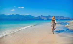 Kobieta na rowerze przemierza piaszczystą plażę z widokiem na turkusowe morze i góry. To idylliczny krajobraz, jak z marzeń o podróży na półwysep Indochiński.