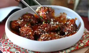 Chrupiący kurczak gochujang w sosie, posypany sezamem i orzeszkami, podany w białej misce.