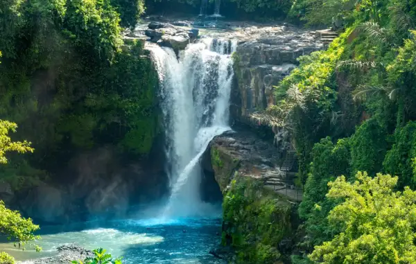 Wodospady na Bali, otoczone bujną zielenią, z kaskadami wody wpadającymi do turkusowej rzeki. Schody prowadzą do punktu widokowego.